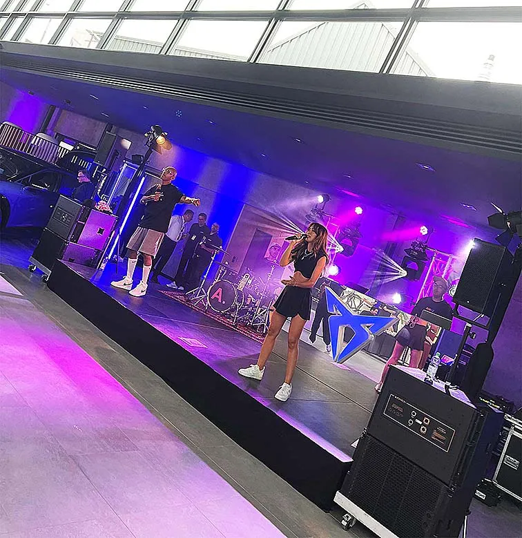 Zwei Personen singen und tanzen auf einer Podestfläche, umgeben von Lautsprechern und Scheinwerfern, auf einer Bühne in einem Autohaus